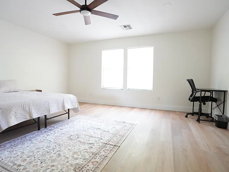 Spacious bedroom with light wood floors, ceiling fan, and dual windows, plus a dedicated desk area for work or study.