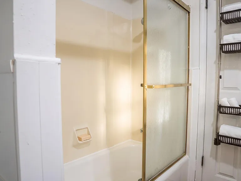 Full bath with tub/shower combo, sliding glass door, and convenient storage shelves for towels and toiletries.