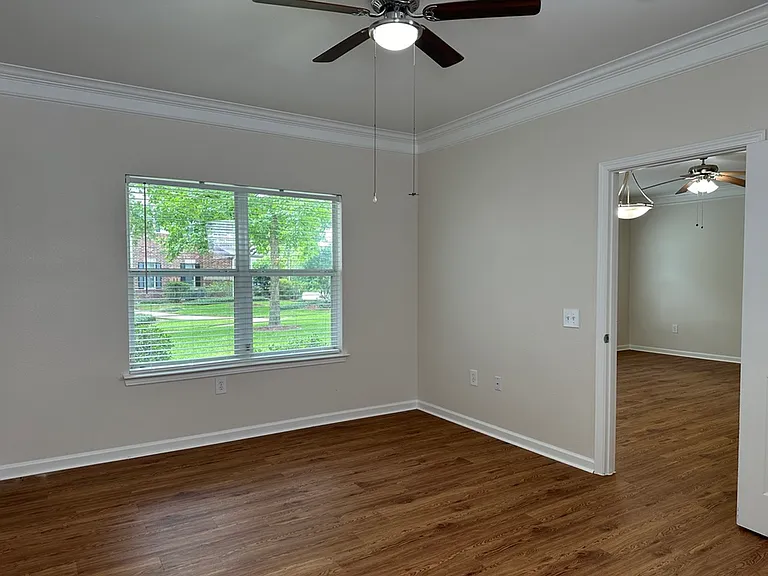 Bright and spacious room featuring elegant wood-style flooring, ceiling fan, and a large window offering lush green views. Bright and spacious room featuring elegant wood-style flooring, ceiling fan, and a large window offering lush green views.
