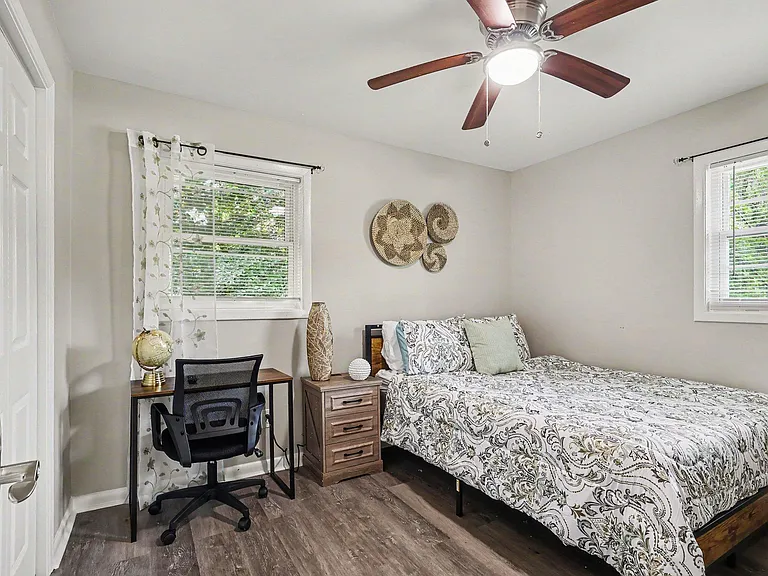 Furnished bedroom with a full-size bed, nightstand, and a dedicated desk area, plus two windows and a ceiling fan.