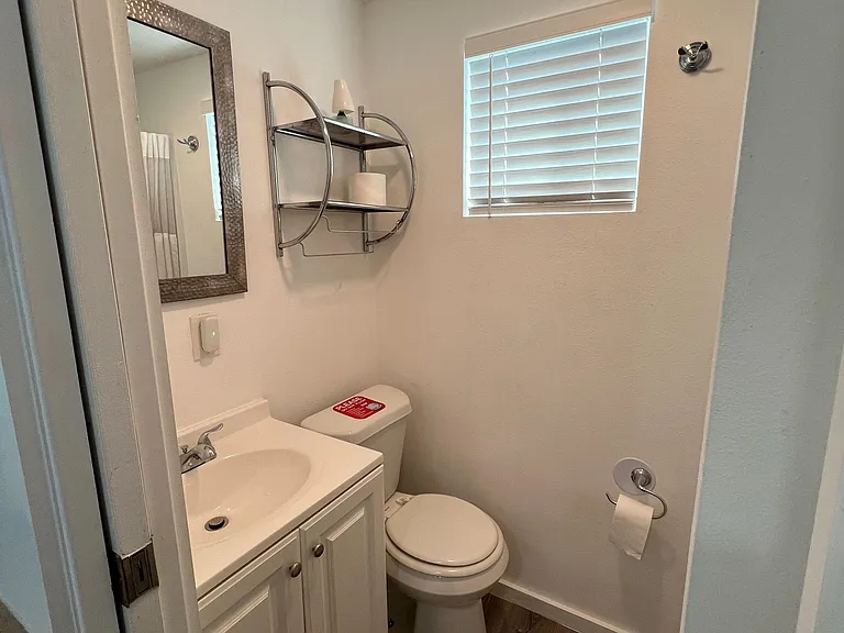 Efficient half bath with a vanity sink, wall mirror, storage shelving, and window with blinds for privacy and light.