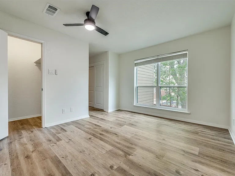 Room featuring wood-finish flooring, a large window with blinds, a black ceiling fan with integrated lighting, and a closet with a bi-fold door