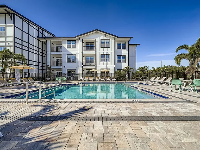 Luxurious apartment complex with a pristine outdoor pool area, surrounded by stylish loungers and palm trees under a clear blue sky.