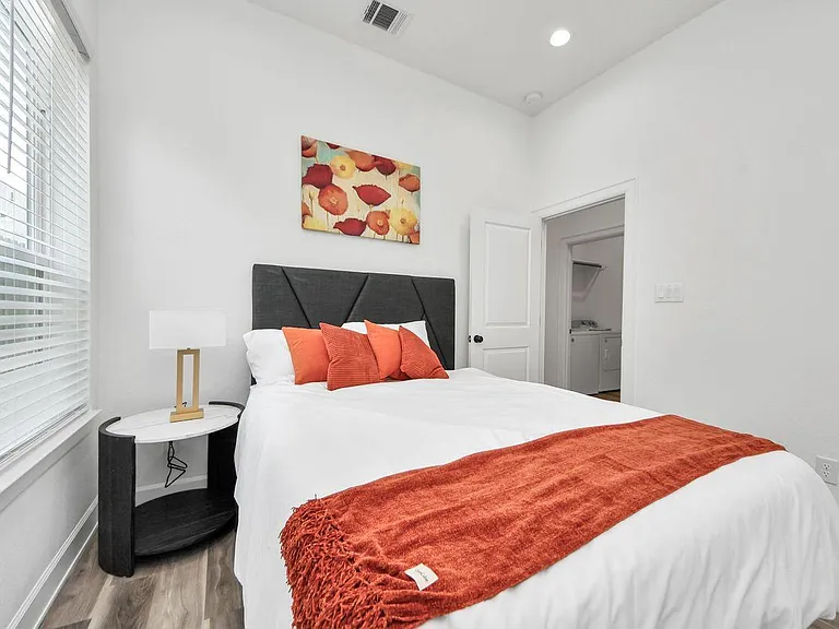 Furnished bedroom with a comfortable bed, large window with blinds, and convenient access to nearby laundry.