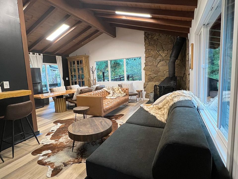 Open-concept living room with vaulted wood-beam ceilings, stone fireplace, and large windows. Cozy seating and rustic-modern decor create the perfect mountain retreat for relaxing or entertaining.