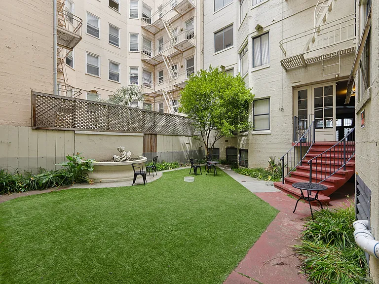 Courtyard with a wooden fence, with a small garden featuring a fountain and a statue, and a staircase that leads up the the entrance of the apartment building