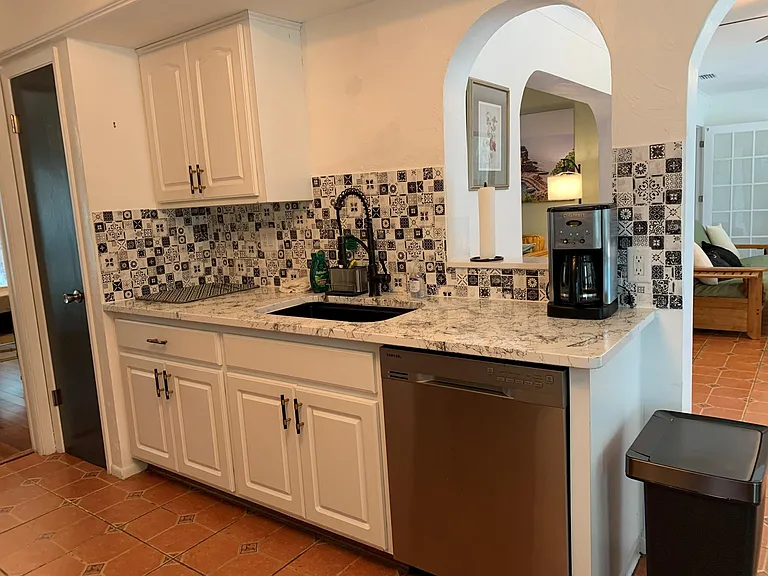 One side of the kitchen with a pantry closet with a door, counter, sink, cabinets, dishwasher, coffee maker, a roll of paper towel on a holder, and a trash can. The pantry and cabinets are full of supplies.
