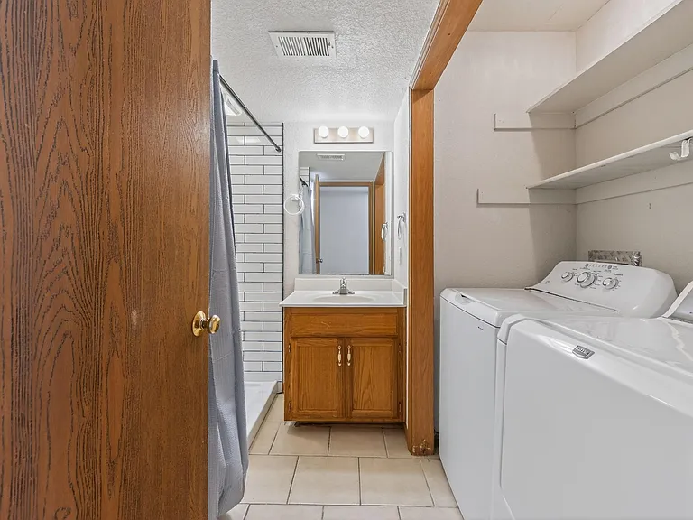 Full bathroom with tub/shower combo, vanity storage, and in-room washer/dryer with adjacent shelving for linens.