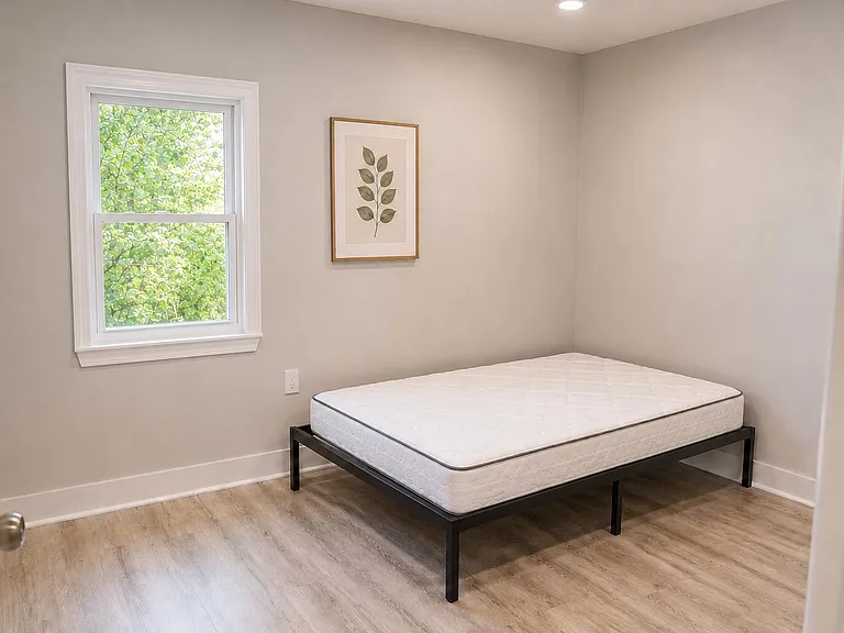 Simple bedroom with a bed frame and mattress, one window with leafy view, neutral walls, and wood-look flooring.