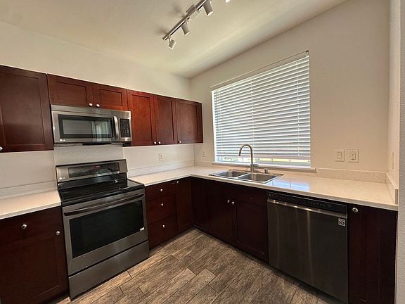 Modern kitchen featuring stainless steel appliances, rich wood cabinetry, and quartz countertops. The space includes a double-basin sink under a large window, track lighting, and sleek tile flooring_offering a clean and functional layout perfect for every