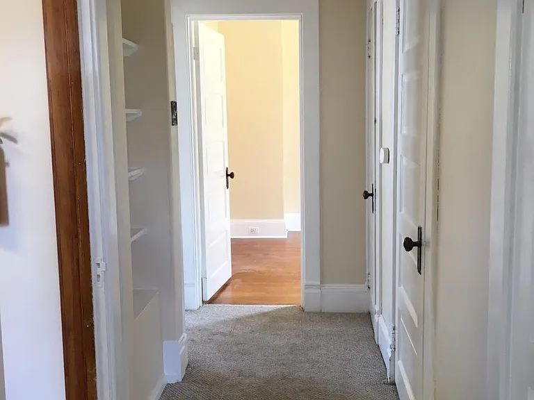 Hallway view from Right bedroom. Kitchen doorway, built in shelves, and living room doorway on the left. Other bedroom straight ahead. Bathroom and hallway closet door on the right.