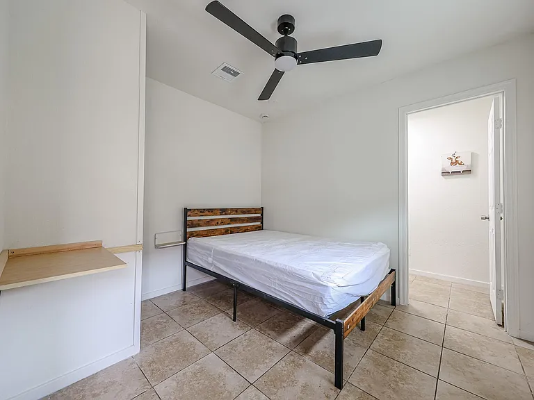 Simple bedroom with tile flooring, ceiling fan, and bed frame, plus a built-in desk/shelf area for work or study.