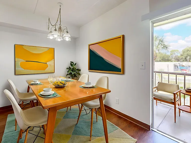 Bright and inviting dining area with modern decor, featuring a wooden table set for four, vibrant abstract artwork, and a petite balcony view.