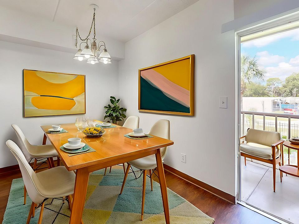 Bright and inviting dining area with modern decor, featuring a wooden table set for four, vibrant abstract artwork, and a petite balcony view.