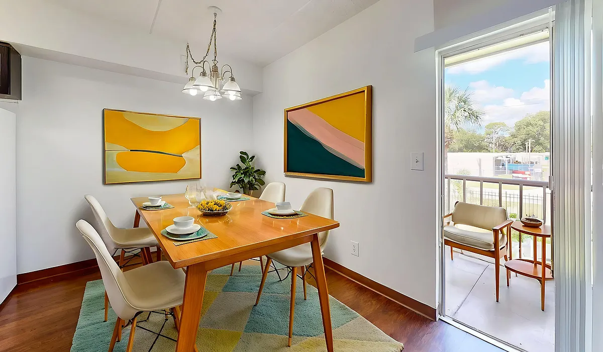 Bright and inviting dining area with modern decor, featuring a wooden table set for four, vibrant abstract artwork, and a petite balcony view. - Windsor Manor