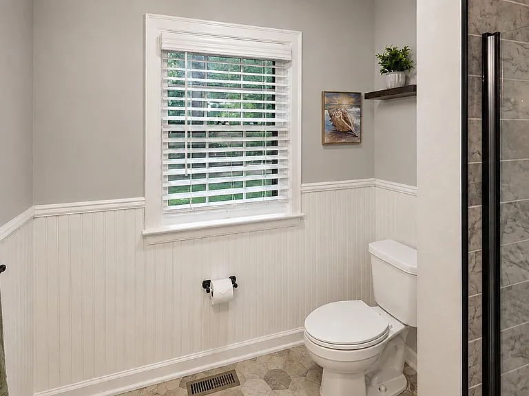 Clean full bath with a window for natural light, beadboard wainscoting, and a tiled shower with glass door.