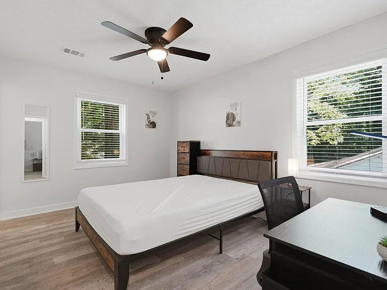 Spacious bedroom with a ceiling fan, large windows with blinds, and room for a work desk alongside the bed.
