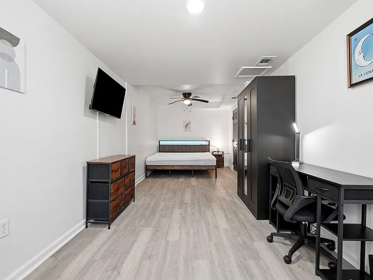 Spacious furnished bedroom with a bed, wardrobe storage, dresser, wall-mounted TV, and a dedicated work desk.