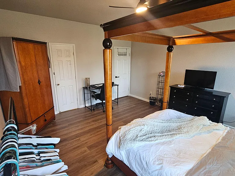 Spacious furnished bedroom with a four-poster bed, dresser with TV, work desk, and wood-look flooring for easy upkeep.