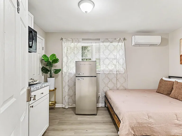 Private bedroom with a full bed, window with blinds and curtains, in-room mini fridge, and wall-mounted A/C for comfort.