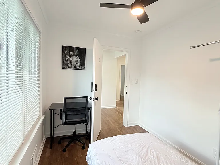 Private bedroom with hardwood floors, ceiling fan, large window with blinds, and a desk nook for work or study.