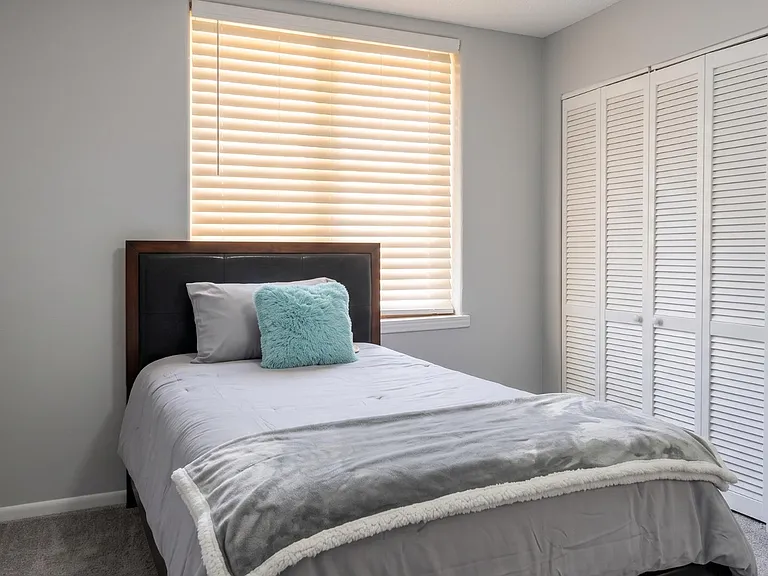 Furnished bedroom with a comfortable full bed, window with blinds, and a wide closet with louvered doors for storage.