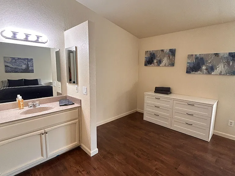 Spacious bedroom with wood-style flooring, dresser storage, and an open vanity area with large mirror and sink.