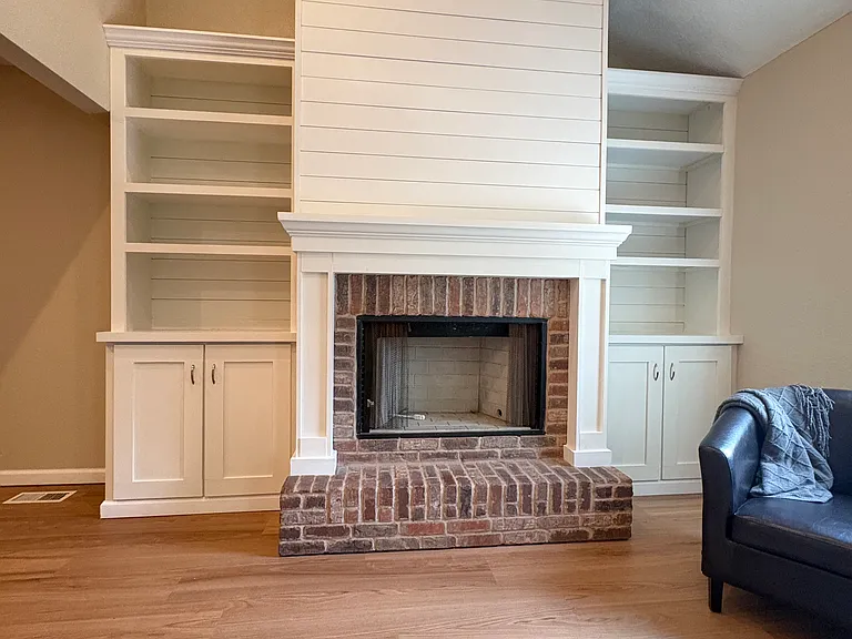 Features a brick hearth surrounded by a custom-built mantel, built-in cabinetry, and upper display shelves. The fireplace is decorative and provided for aesthetic purposes only.