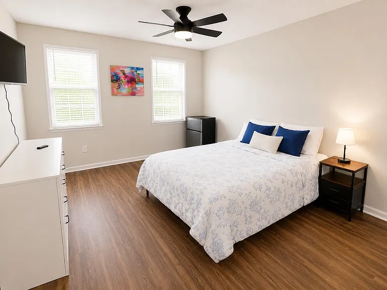 Spacious bedroom with two windows, a ceiling fan, wall-mounted TV, mini fridge, and dresser plus bedside table.