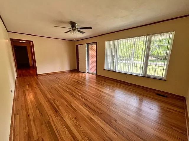 Spacious living room featuring beautiful hardwood floors, neutral wall colors, and abundant natural light from large front windows with vertical blinds. The open layout provides plenty of room for multiple furniture arrangements and entertaining. Includes