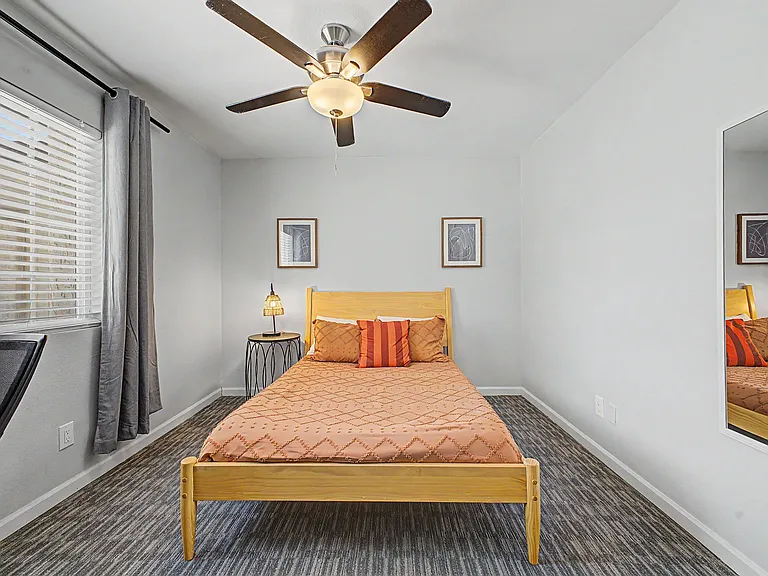 Private bedroom with ceiling fan, large window with blinds and curtains, carpeted floors, and full-length mirror.