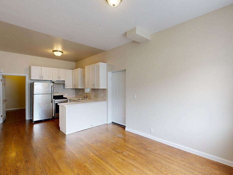 Bright, empty kitchen with hardwood floors, cabinets, and stainless steel appliances, including a fridge and stove. Neutral walls and ample lighting.