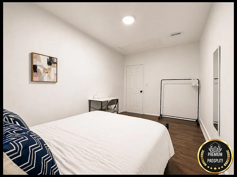 Private bedroom with a full-size bed, desk workspace, and clothing rack, finished with wood-style flooring.