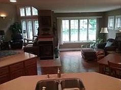 Center kitchen island overlooks triple fireplace and Hearth Room with tall window adorned by tantrum windows for full view of woods