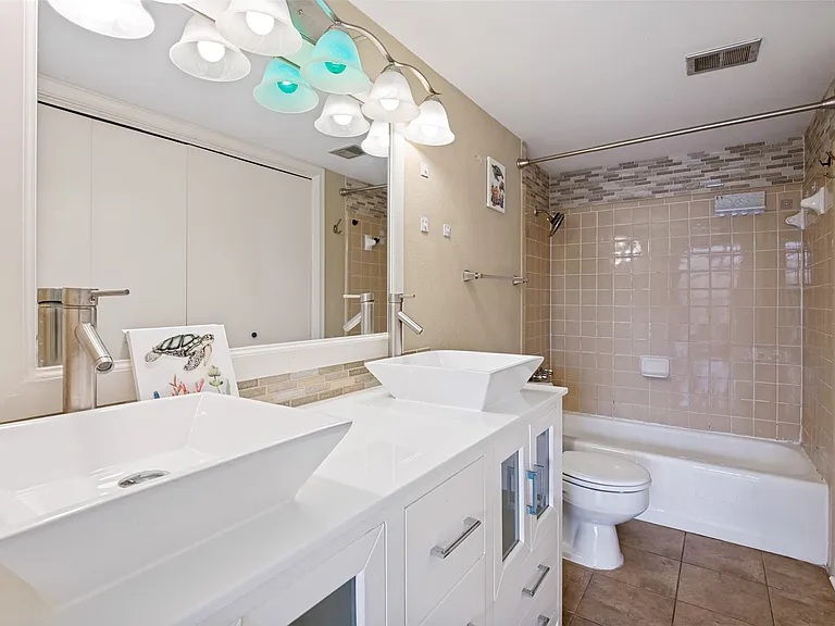 Dual vessel sinks with modern faucets, white vanity with cabinet storage, full vanity mirror, and a tiled shower/tub combination