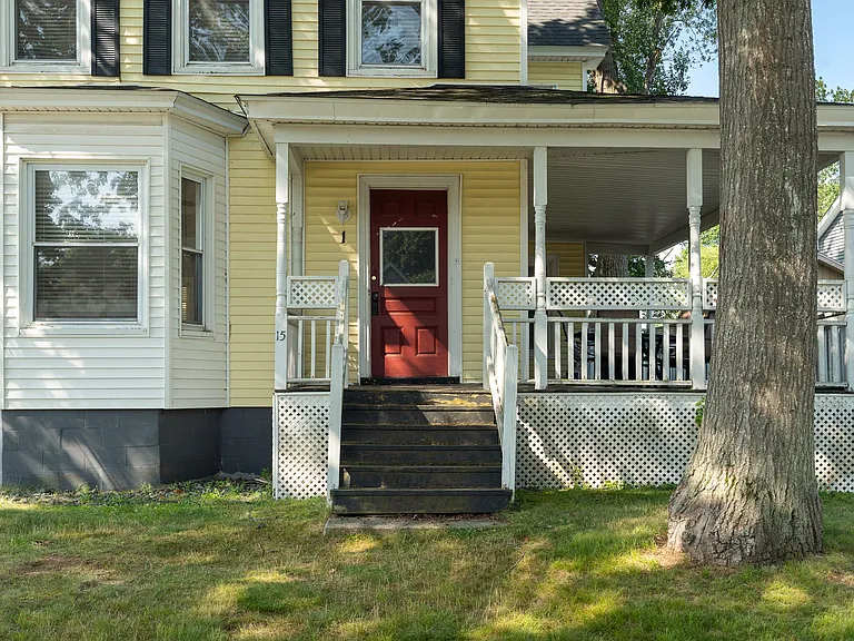 Welcome to this charming, traditional home with a large, inviting front porch. The exterior is painted a cheerful yellow with crisp white trim and black shutters. A bright red front door stands out, framed by a classic white porch railing. Dark stairs lea