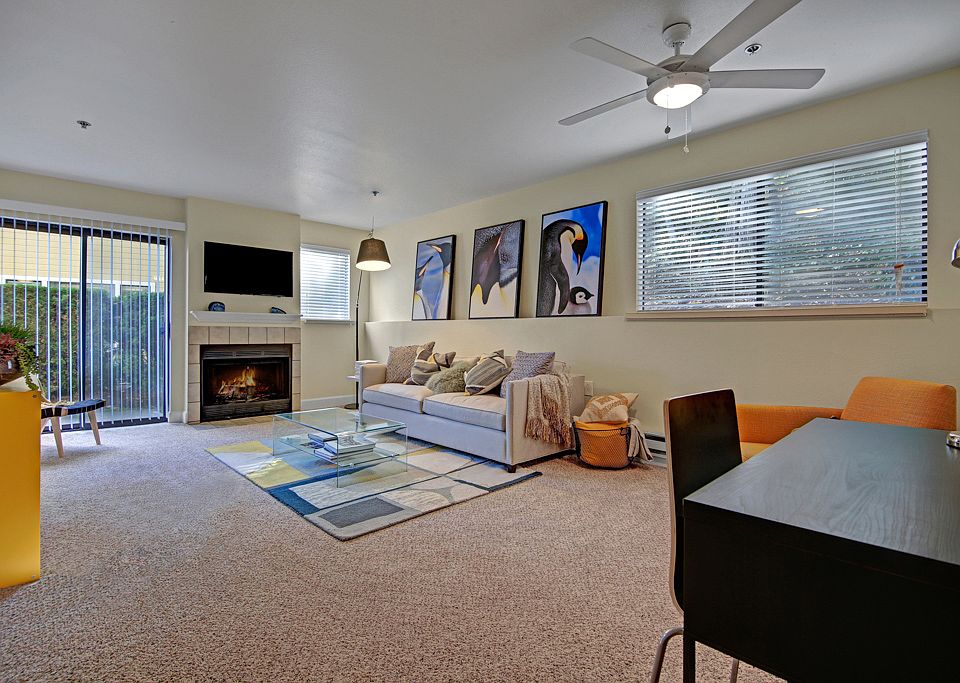 Living room with couch, desk, armchair, large windows, ceiling fan, fireplace, and sliding glass door out to a patio