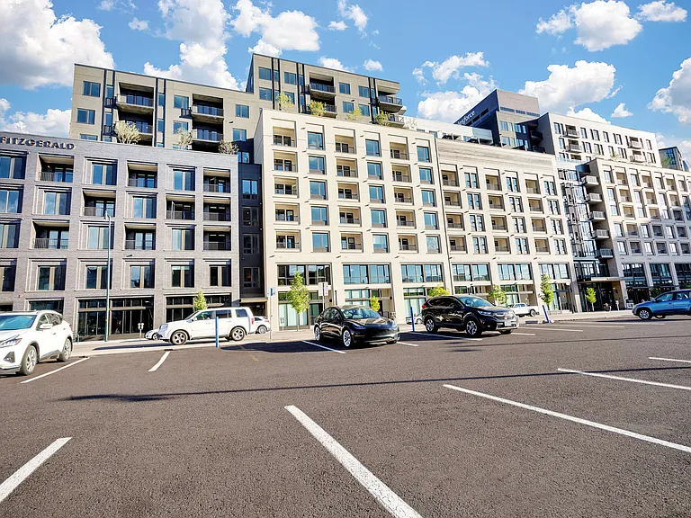 Modern urban apartment complex with sleek architecture, large windows, and ample parking, set against a bright blue sky.