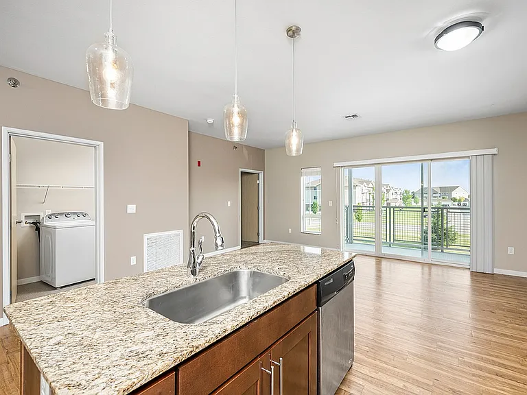 Bright apartment interior featuring a modern granite countertop island, stylish pendant lighting, open-concept living area, and large sliding glass doors leading to a balcony with scenic views. Bright apartment interior featuring a modern granite countert