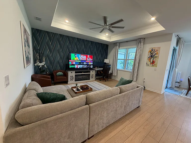 Spacious living room with tray ceiling, ceiling fan, large window, and ample seating anchored by an entertainment console.