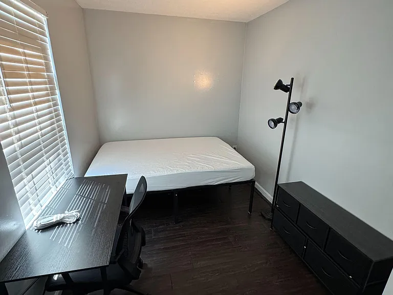 Furnished bedroom with a bed, desk and chair, dresser storage, and a large window with blinds for natural light.