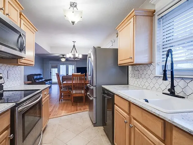 Galley kitchen featuring light wood cabinetry, stone-finish countertops, stainless steel appliances, and a geometric tile backsplash