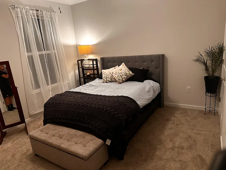 Furnished bedroom with a queen bed, upholstered headboard, bedside lighting, and a window with sheer curtains.