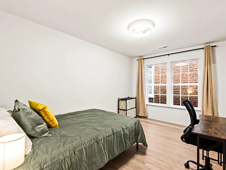 Furnished bedroom with a full bed, work desk, and large window with curtains for comfortable daily living.