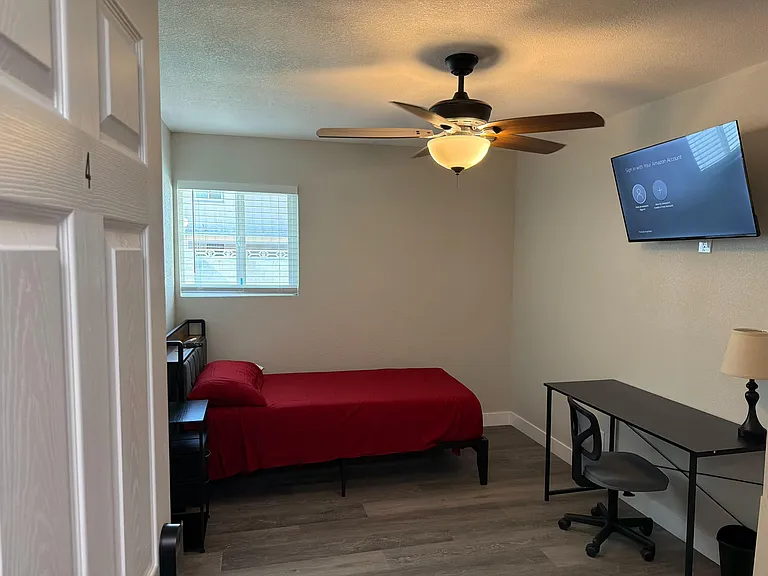 Furnished bedroom featuring a ceiling fan with light, window blinds, wall-mounted TV, and a dedicated desk workspace.
