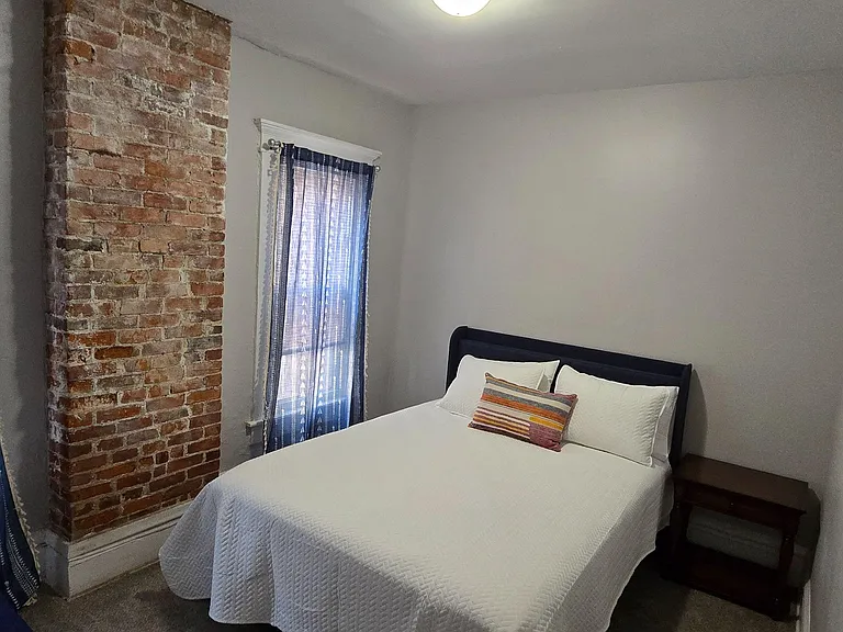 Furnished bedroom with exposed brick accent, two windows with curtains, and a bed with side table for easy everyday living.