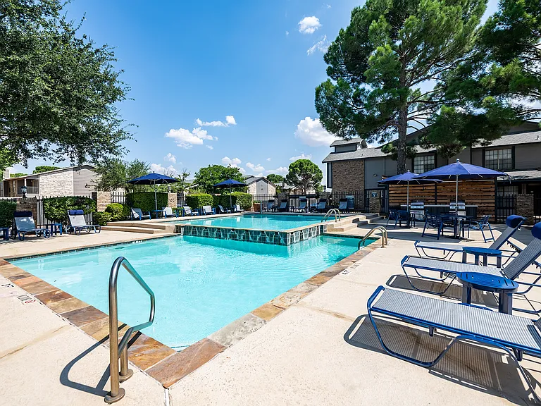 A swimming pool and spa are shown on a sunny day next to a sundeck with chaise lounge chairs and picnic tables with umbrellas.