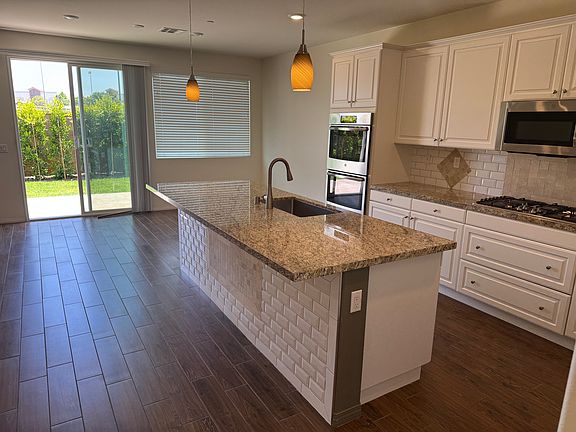 View of kitchen with sink in kitchen island and pendant lights in foreground, double oven, range and above range microwave, and dining area in background, along with sliding glass door in backyard.