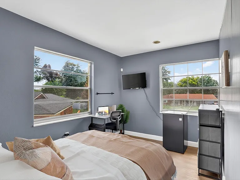 Bedroom offers two large windows, a work desk setup, wall-mounted TV, and space for a mini fridge and storage.