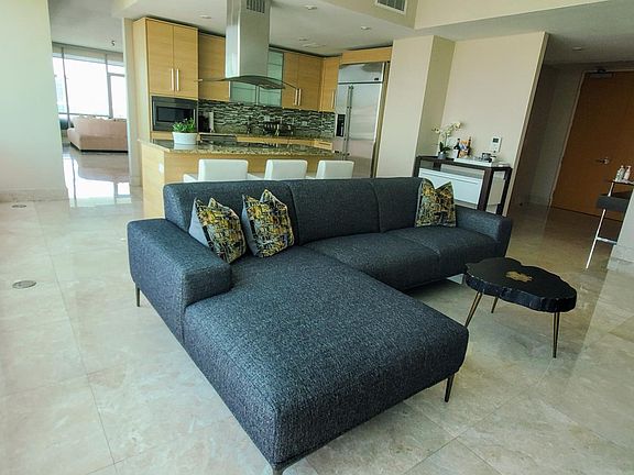 Living room with open kitchen in the background. Can glimpse the family room in the back and to the right side can see the entryway to the apartment.